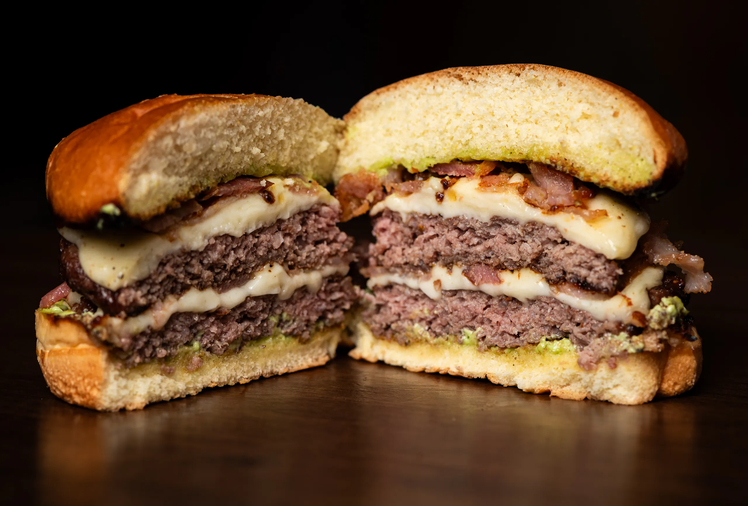 Brouss Duplo Bacon cortado ao meio: duas carnes bovinas, queijo derretido entre elas, bacon e maionese verde sobre o pão selado.